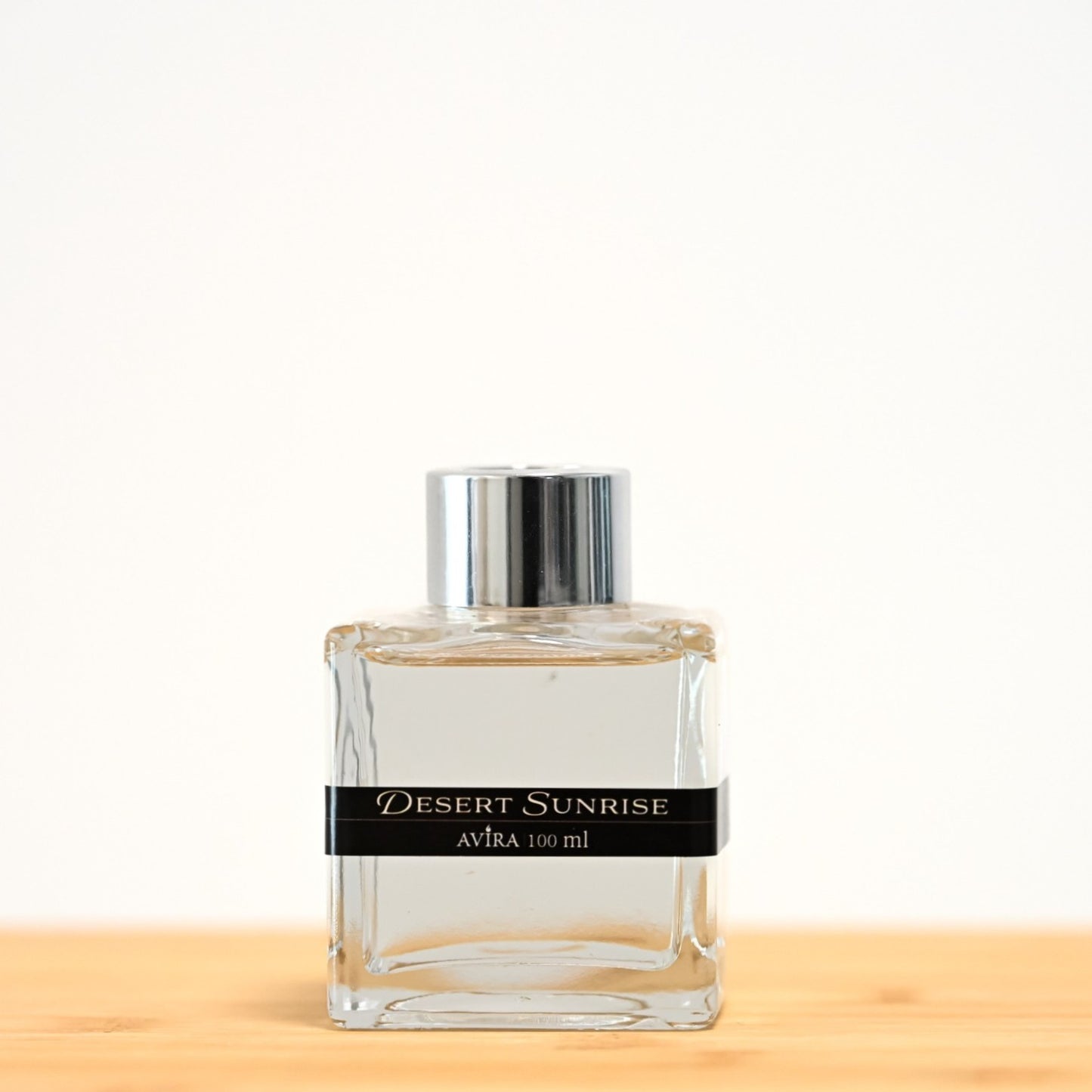 מפיץ ריח 100 מ"ל בניחוח זריחה במדבר, בכלי זכוכית שקוף בצורת קובייה – אטום בפקק ומלווה בקולר כסוף – ובחזית מדבקת פס שחורה.