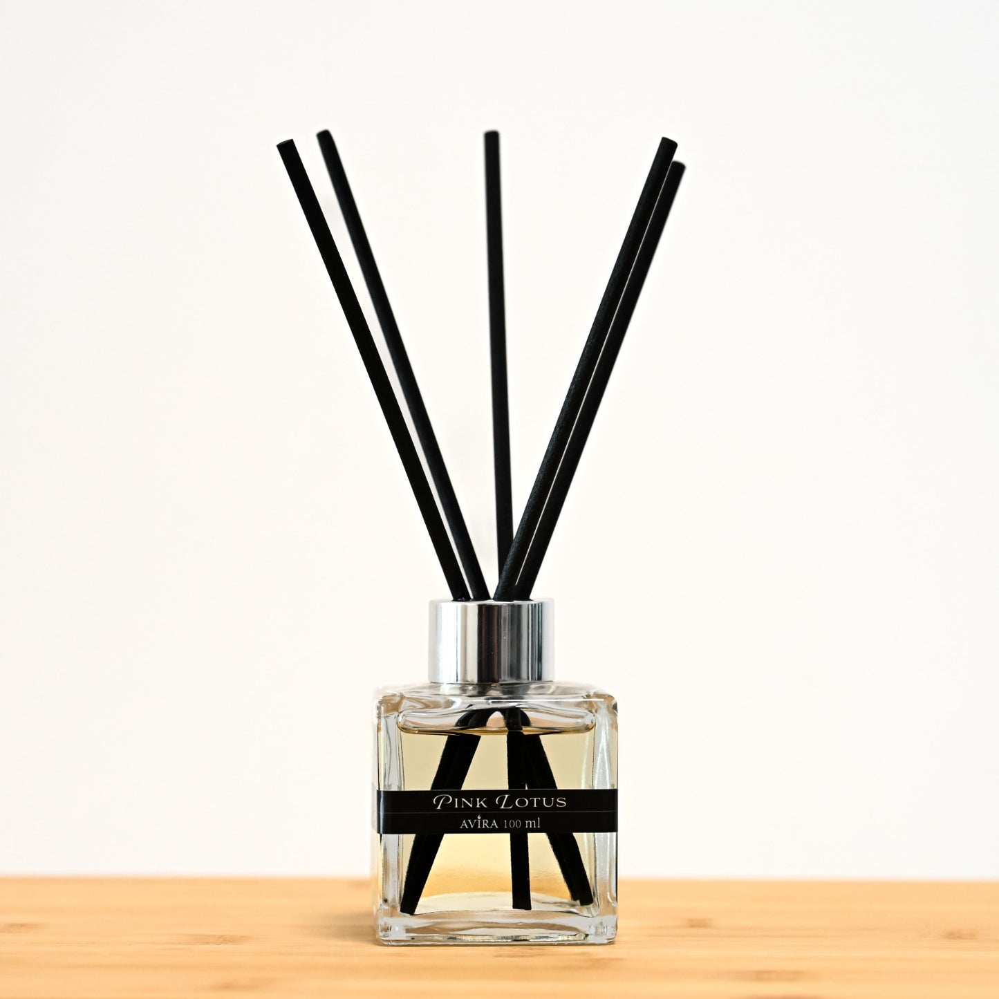 מפיץ ריח 100 מ"ל בניחוח לוטוס ורוד עם מקלות שחורים, בכלי זכוכית שקוף בצורת קובייה – אטום בפקק ומלווה בקולר כסוף – ובחזית מדבקת פס שחורה.