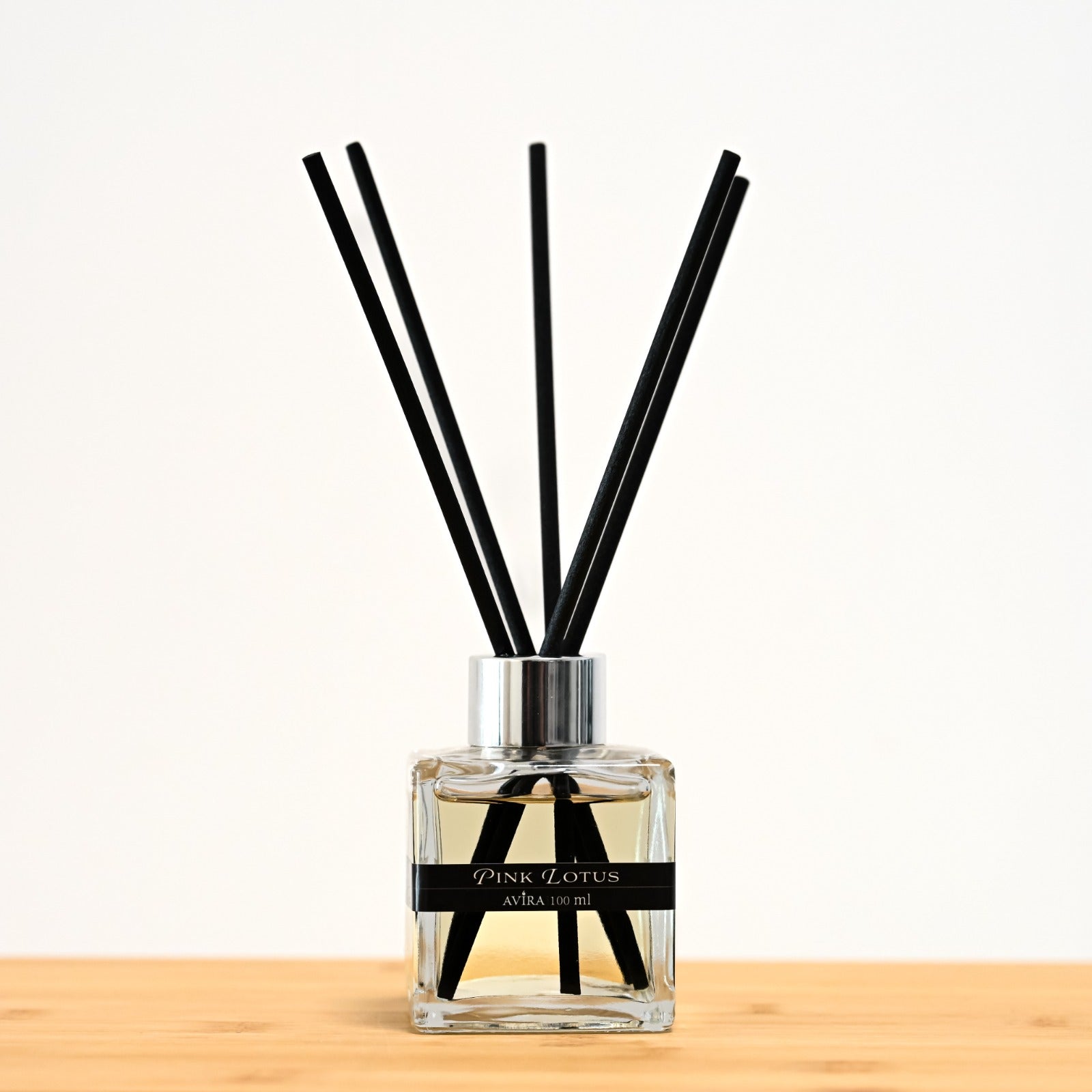 מפיץ ריח 100 מ"ל בניחוח לוטוס ורוד עם מקלות שחורים, בכלי זכוכית שקוף בצורת קובייה – אטום בפקק ומלווה בקולר כסוף – ובחזית מדבקת פס שחורה.