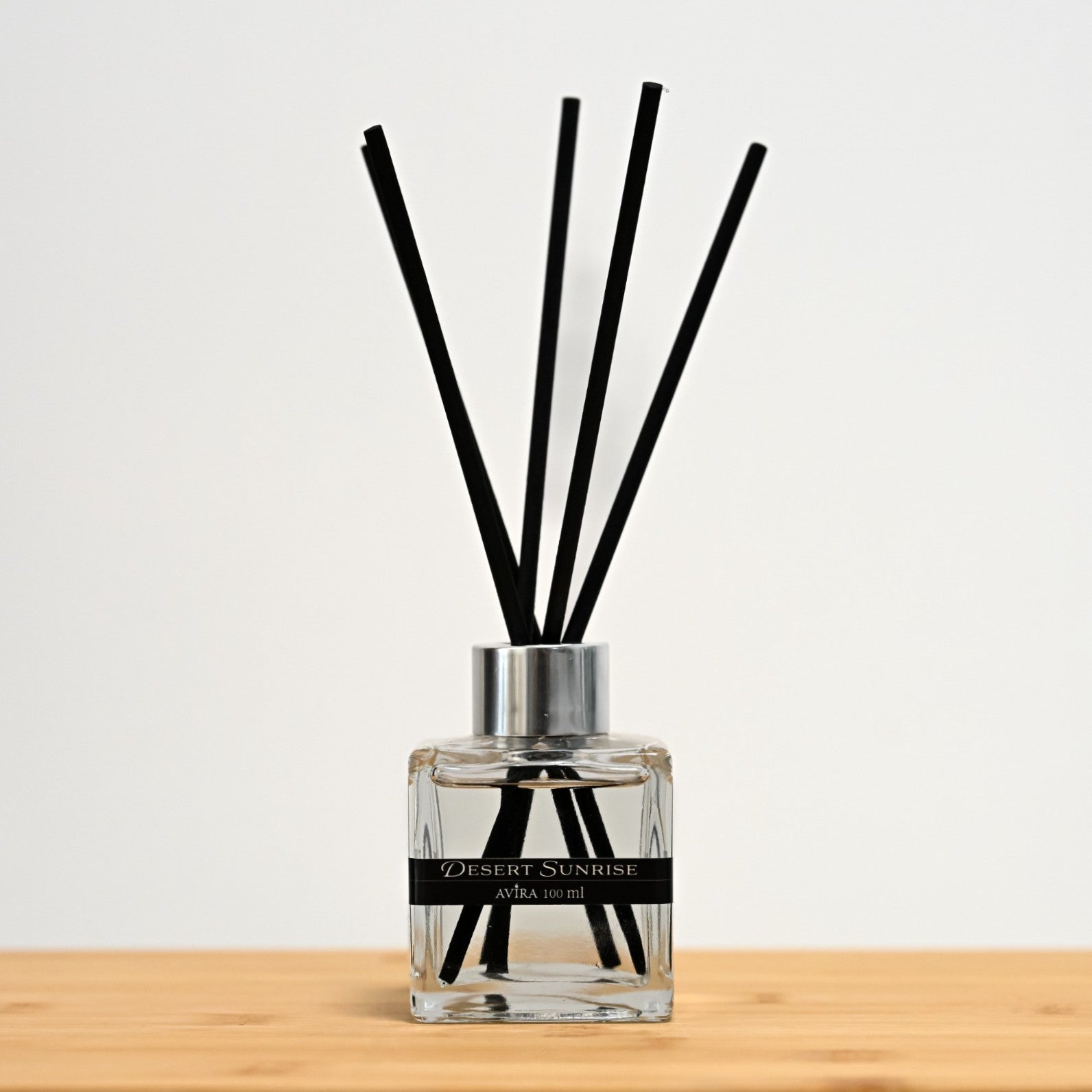 מפיץ ריח 100 מ"ל בניחוח זריחה במדבר עם מקלות שחורים, בכלי זכוכית שקוף בצורת קובייה – אטום בפקק ומלווה בקולר כסוף – ובחזית מדבקת פס שחורה.