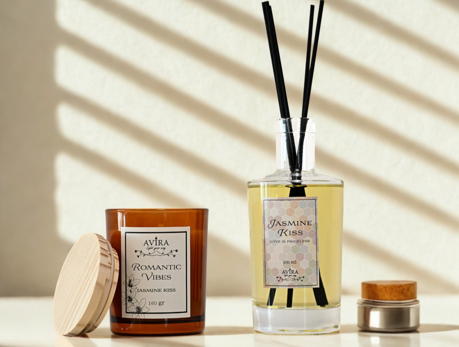 נר בכוס אמבר חומה עם מכסה עץ ומדבקה לבנה שעליה כתוב "ROMANTIC VIBES", ומימינו מפיץ ריח שקוף עם מקלות שחורים בניחוח יסמין
